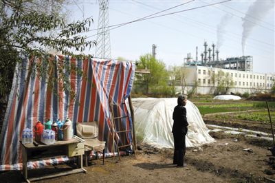 张占权老伴站在他们临时的住所前,不远处是陪伴了他们半辈子的化工厂。